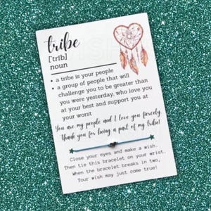 May include: A white rectangular card featuring a dreamcatcher illustration and the definition of the word tribe. The card includes a thin blue string bracelet with a small heart charm and printed text explaining a wish-making tradition.