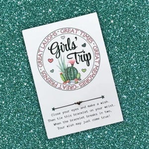 May include: A white card with the words "Girls' Trip" in a circular design with cacti and hearts. The card includes instructions to tie a bracelet and make a wish. The background is a sparkly teal color.