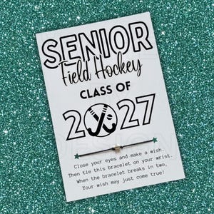 May include: A white rectangular card featuring the black text SENIOR Field Hockey CLASS OF 2027 with a hockey stick graphic inside the zero. A thin black string bracelet with a small star charm is attached to the card, which includes wish instructions.