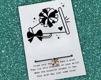 May include: A black string bracelet with a small gold-colored bow charm attached to a white card. The card features a black graphic of a megaphone, a pom-pom, and a bow, with the text: Close your eyes and make a wish. Then tie this bracelet on your wrist. When the bracelet breaks in two, Your wish may just come true!