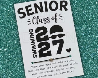 May include: A black string bracelet with a small silver star charm attached to a white card. The card features black text reading SENIOR Class of 2027 SWIMMING, a swimming cap and goggles icon, a heart, and instructions to make a wish.