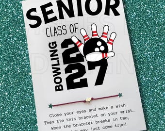 May include: A white card featuring the text SENIOR CLASS OF 27 with a graphic of a bowling ball and pins. A thin maroon string bracelet with a small silver heart charm is attached to the card, which includes printed instructions for a wish.