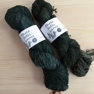 May include: Two skeins of dark green yarn with white labels that read "Three fates yarns". The yarn is tightly wound and appears soft. The background is a light wood grain.