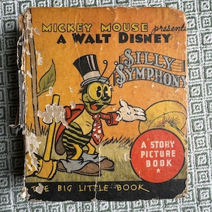 Puede incluir: Una portada de libro vintage con Mickey Mouse presentando una Silly Symphony de Walt Disney. La portada presenta una ilustración de dibujos animados de un insecto verde con un sombrero de copa y una corbata de lazo, sosteniendo una escoba. El texto en la portada dice "Mickey Mouse presenta Una Silly Symphony de Walt Disney Un libro de imágenes de historia El gran libro pequeño".