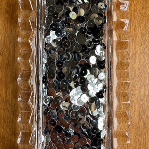 May include: A rectangular clear glass dish filled with shiny silver sequins. The dish has a decorative, scalloped edge. The sequins are round and reflect light, creating a shimmering effect. The dish is on a wooden surface.