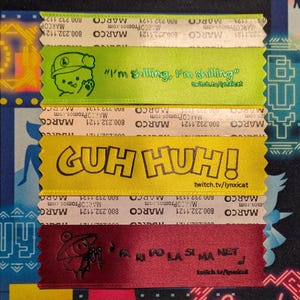 Könnte beinhalten: Nahaufnahme von vier farbigen Bändern mit Text und Cartoon-Grafiken. Die Bänder sind grün, gelb und rot. Das grüne Band sagt "I'm shilling, I'm shilling" mit einer Cartoon-Figur. Das gelbe Band sagt "GUH HUH!"