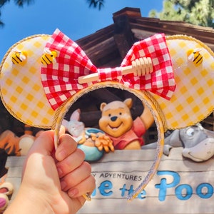 May include: Yellow and red Minnie Mouse ears with a gingham bow and a honey pot charm. The ears are decorated with small yellow bees. The ears are in front of a sign that says "Winnie the Pooh Adventures".