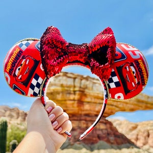 May include: Red and black sequined Minnie Mouse ears with a Cars theme. The ears are decorated with Lightning McQueen and the text "Lightning McQueen".