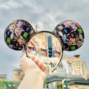 May include: A headband featuring two circular ears covered in a colorful, repeating pattern of various cartoon characters, including a small tree-like figure, with a metallic brown braided trim along the edges of the ears and the headband frame.