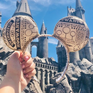 May include: A brown and gold Mickey Mouse ear headband with a printed design featuring the words "Quidditch at Hogwarts" and "Gwarts". The headband is in front of a building with a castle-like architecture.