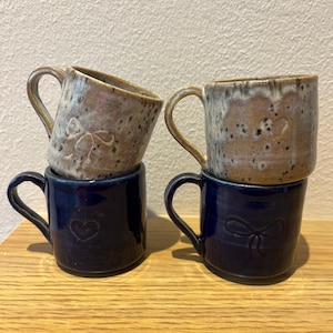 May include: Four ceramic mugs arranged in two stacks. The top two mugs feature a speckled beige and brown glaze with embossed bow designs. The bottom two mugs are solid dark blue with embossed heart and bow motifs on their front surfaces.