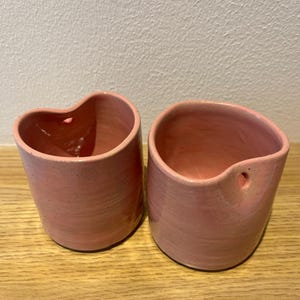 May include: Two cylindrical ceramic vessels in a soft pink speckled glaze. Each container features an irregular, organic rim shape and a single small circular hole near the top edge, designed for hanging or mounting purposes on a wall surface.