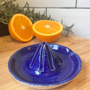 May include: A vibrant cobalt blue ceramic citrus juicer with a ridged reamer and a shallow bowl. Two halves of an orange are in the background. The juicer sits on a wooden surface, with a white tiled wall and greenery behind.
