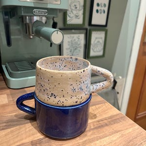 May include: Two-tone ceramic mugs. The top mug is beige with black speckles, and the bottom mug is a deep blue. The mugs are stacked on top of each other. The mugs are on a wooden surface, with a mint green espresso machine in the background.