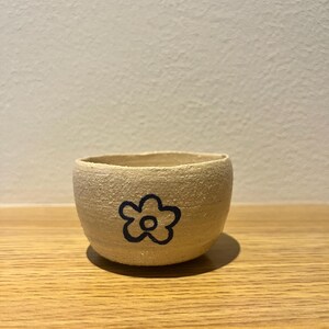 May include: A small, beige ceramic bowl with a black flower design. The bowl has a rounded shape and a textured surface. The flower is a simple outline. The bowl is sitting on a wooden surface.