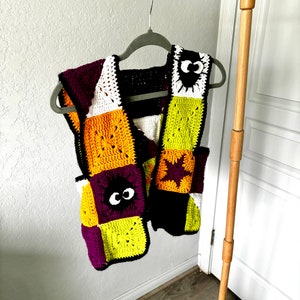 May include: A crocheted vest with a patchwork design featuring squares in purple, white, yellow, orange, black, and green. The vest has a black and white eye design on two of the squares.