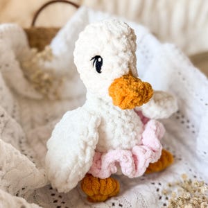 May include: A white crocheted duck plush toy wearing a pink ruffled skirt. The duck has orange yarn for its beak and feet.