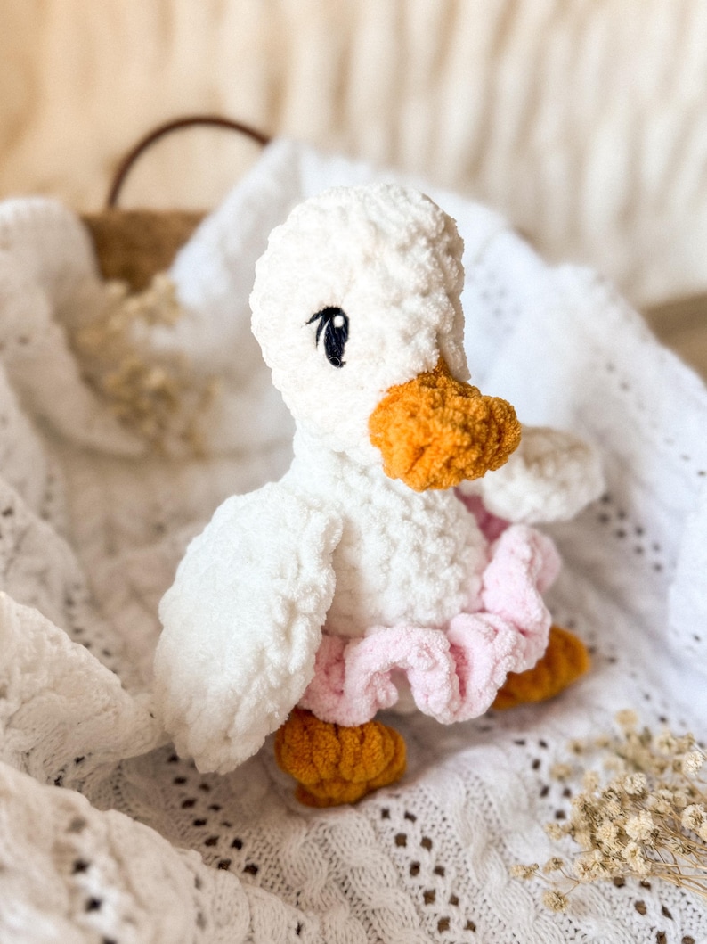 May include: A white crocheted duck plush toy wearing a pink ruffled skirt. The duck has orange yarn for its beak and feet.