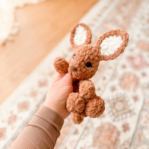 K&ouml;nnte beinhalten: Ein handgeh&auml;kelter brauner H&auml;schen mit wei&szlig;en Innenohren und schwarzen Augen. Das Pl&uuml;schtier hat ein strukturiertes, weiches Aussehen. Das H&auml;schen wird vor einem gemusterten Teppich gehalten.