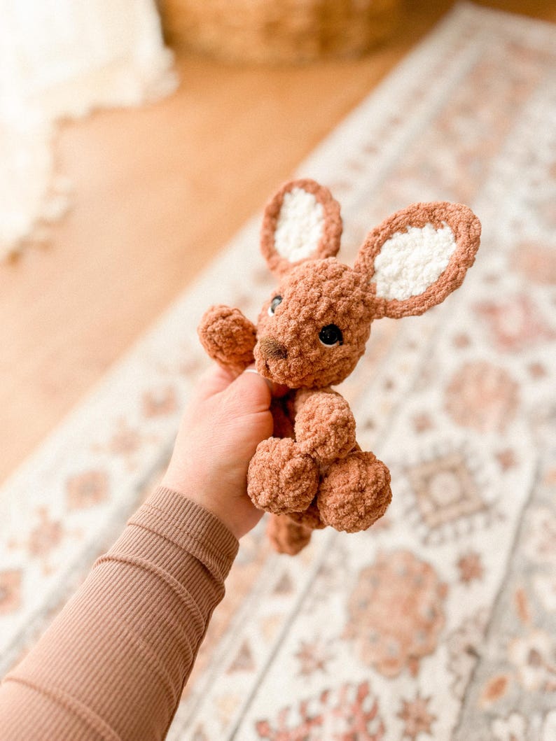 K&ouml;nnte beinhalten: Ein handgeh&auml;kelter brauner H&auml;schen mit wei&szlig;en Innenohren und schwarzen Augen. Das Pl&uuml;schtier hat ein strukturiertes, weiches Aussehen. Das H&auml;schen wird vor einem gemusterten Teppich gehalten.