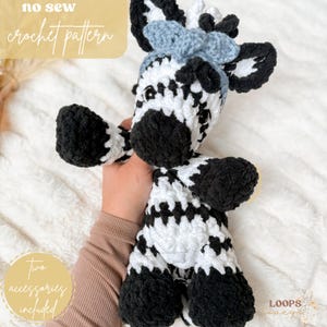 Könnte beinhalten: Handgehäkeltes Zebra-Plüschtier in Schwarz und Weiß mit einer blauen Schleife. Das Plüschtier hat schwarze Hufe, eine schwarze Nase und einen schwarz-weiß gestreiften Körper. Der Text "no sew crochet pattern" und "Two accessories included" sind sichtbar.