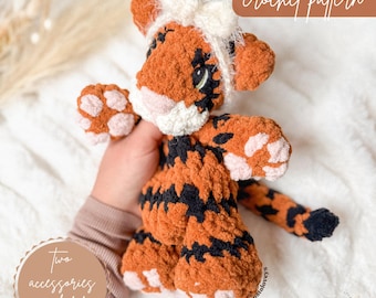 May include: A crocheted tiger toy in shades of orange and black, with white and pink details. The image includes the text "no sew crochet pattern" and "Two accessories included". The toy has a white bow on its head.