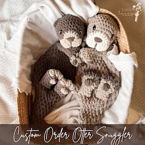 Könnte beinhalten: Zwei handgefertigte Otter-Kuscheltiere aus Häkelarbeit in einem Weidenkorb. Einer ist braun, der andere hellbraun. Beide haben hellbraune Gesichter und Pfoten. Der Text "Custom Order Otter Snuggler" befindet sich am unteren Bildrand.