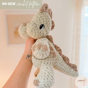May include: A handmade crochet dinosaur in shades of beige and brown. The plush toy features a textured, fluffy design with a detailed head, small black eyes, and a row of brown spikes down its back. The text "NO SEW crochet pattern" is at the top.