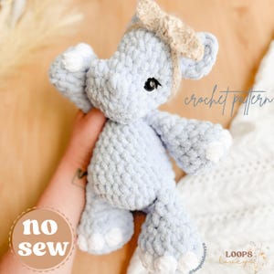 May include: A light blue crocheted elephant plush toy with white accents on its feet and ears. The toy has a beige bow and black eyes. The words "crochet pattern" and "no sew" are visible. The toy is handmade.