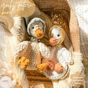 Może przedstawiać: Dwie pluszowe zabawki amigurumi w kształcie gęsi, jedna biała z żółtym dziobem i jasnozielonym kapeluszem, a druga biała z żółtym dziobem i jasnoniebieskim kapeluszem. Zabawki leżą w wiklinowym koszu z beżowym kocem.