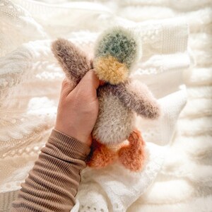 May include: A hand holding a plush toy duck with a green, yellow, and brown patterned head. The duck has orange feet and is sitting on a white knitted blanket.