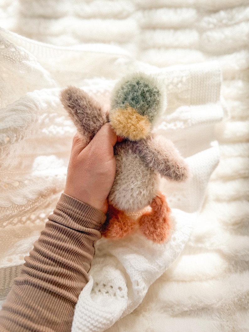 May include: A hand holding a plush toy duck with a green, yellow, and brown patterned head. The duck has orange feet and is sitting on a white knitted blanket.