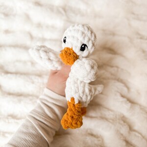 May include: A white crocheted duck plush toy with orange feet and black eyes. The duck is being held by a hand.