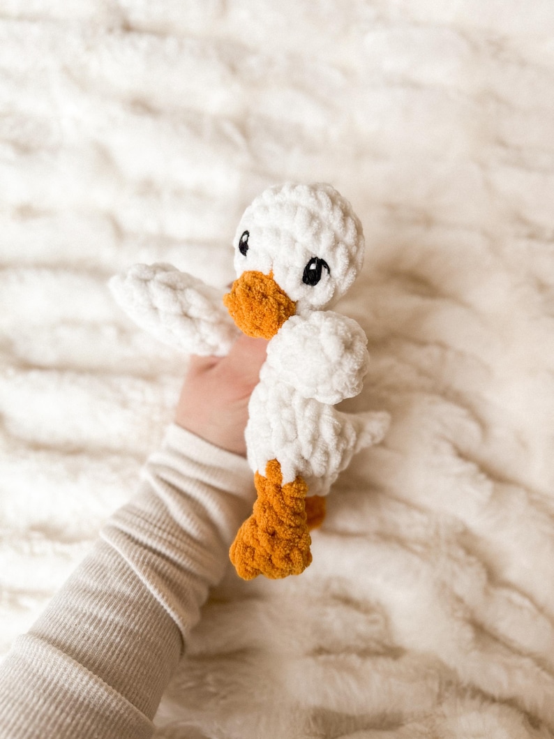 May include: A white crocheted duck plush toy with orange feet and black eyes. The duck is being held by a hand.