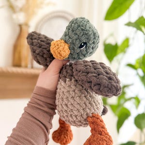 May include: A crocheted plush toy duck with brown, green, and orange yarn. The duck has a small, round, orange beak and black eyes.