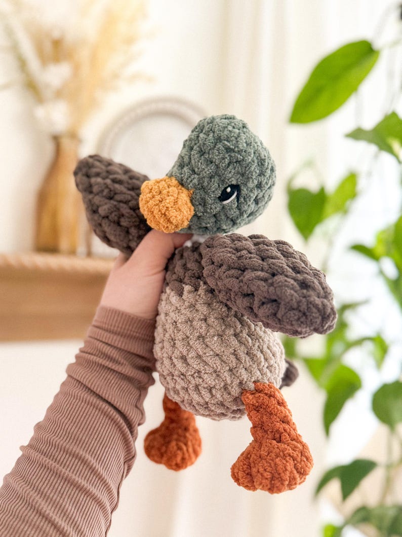 May include: A crocheted plush toy duck with brown, green, and orange yarn. The duck has a small, round, orange beak and black eyes.