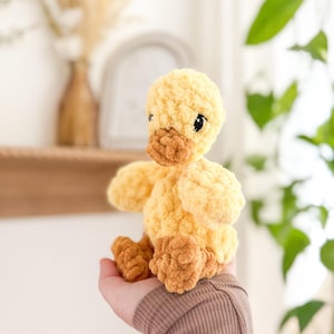 May include: A crocheted yellow duck with brown feet. The duck is sitting in the palm of a hand.