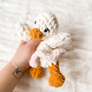 May include: A white crocheted duck plush toy with orange feet and a pink belly. The duck has embroidered eyes and a black beak.