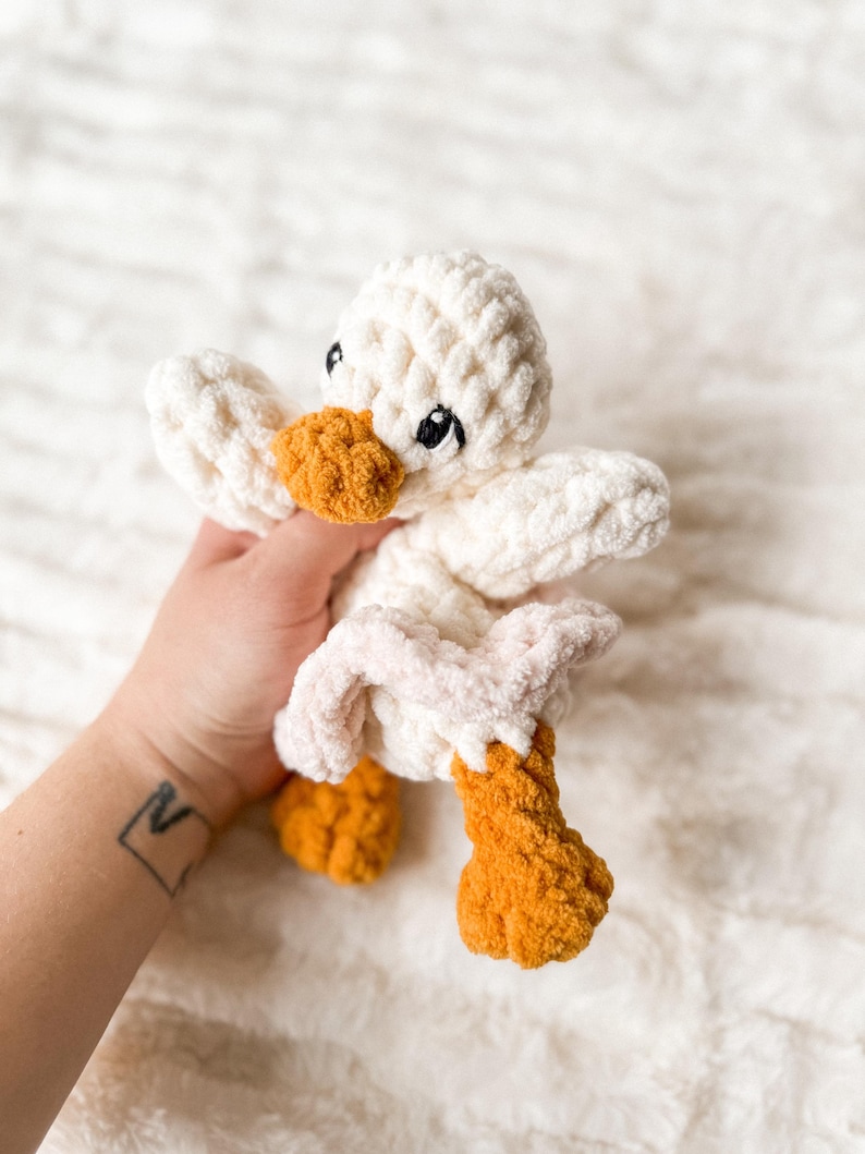 May include: A white crocheted duck plush toy with orange feet and a pink belly. The duck has embroidered eyes and a black beak.