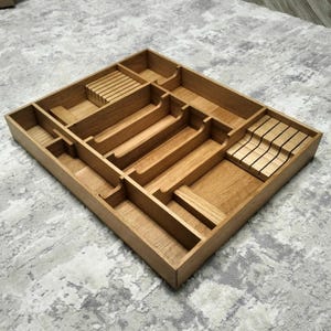 Puede incluir: Organizador de cubiertos de madera con múltiples compartimentos. El organizador es rectangular con varias ranuras y secciones para guardar la cubertería. La madera tiene un color marrón claro natural. Diseñado para cajones.