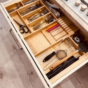 May include: A kitchen drawer with wooden dividers, containing cutlery, utensils, and kitchen tools. Includes forks, spoons, knives, scissors, spatulas, and a potato masher. The drawer is white with a light wood interior.
