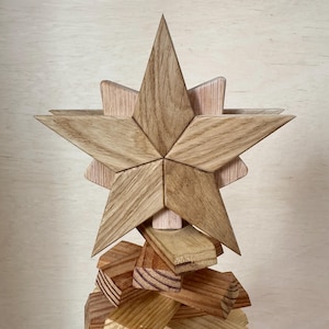 Könnte beinhalten: Ein fünfzackiger Stern aus hellem Holz mit sichtbarer Maserung. Der Stern besteht aus zwei übereinanderliegenden Holzschichten, die einen dreidimensionalen Effekt erzeugen, und ist auf einem Stapel horizontaler Holzblöcke montiert.
