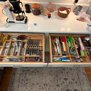 Può includere: Due cassetti da cucina in legno affiancati e aperti, che rivelano scomparti organizzati pieni di posate in acciaio inossidabile, vari utensili da cucina, misurini e assortiti strumenti da cucina disposti in una disposizione ordinata e suddivisa.