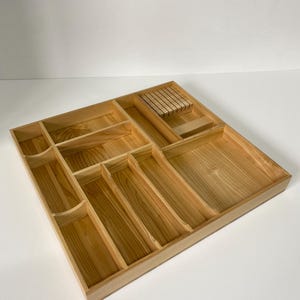 Puede incluir: Una bandeja de cubiertos de madera con múltiples compartimentos para organizar la cubertería y los utensilios de cocina. La bandeja es de madera clara y tiene una sección para cuchillos.