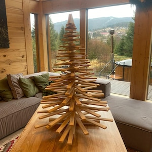 Peut inclure: Une sculpture décorative en forme de sapin de Noël composée de nombreuses lattes de bois horizontales empilées en cône. Les pièces en bois naturel brun clair sont disposées en éventail, se rétrécissant vers une pointe triangulaire au sommet.