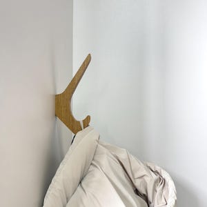 Könnte beinhalten: Ein hellbrauner Wandhaken aus Holz mit geschwungenem, abgewinkeltem Design, der in einer Ecke montiert ist. Der Haken weist eine sichtbare Holzmaserung auf und trägt eine gefaltete, hellbeige Daunenjacke, die am unteren Teil hängt.