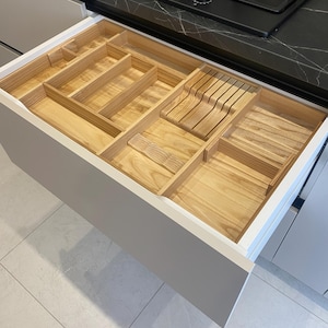 Könnte beinhalten: Geöffnete weiße Küchenschublade mit einem individuellen Holz-Utensilien-Organizer. Der Organizer hat verschiedene Fächer für Messer, Gabeln, Löffel und andere Küchenutensilien. Die Schublade ist geöffnet und zeigt das helle Holz und die organisierte Anordnung.