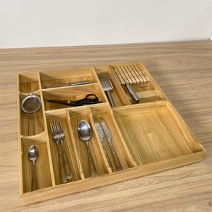 May include: A wooden kitchen utensil organizer with multiple compartments. It contains a variety of kitchen tools, including a whisk, scissors, a grater, a knife block, silverware, and a small strainer. The organizer is made of light-colored wood.