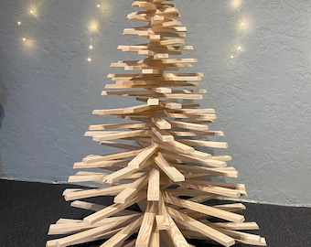 May include: A wooden Christmas tree made from multiple horizontal wooden slats, forming a layered, geometric design. The tree is unpainted, displaying the natural wood grain. It stands on a dark surface against a textured wall.