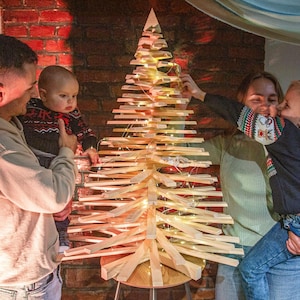 Könnte beinhalten: Ein kegelförmiger Weihnachtsbaum aus gestapelten, horizontalen, hellen Holzlatten. Die Struktur verfügt über eine zentrale vertikale Stütze und wird von dünnen, flexiblen Lichterketten mit warmweißen LED-Leuchten beleuchtet.